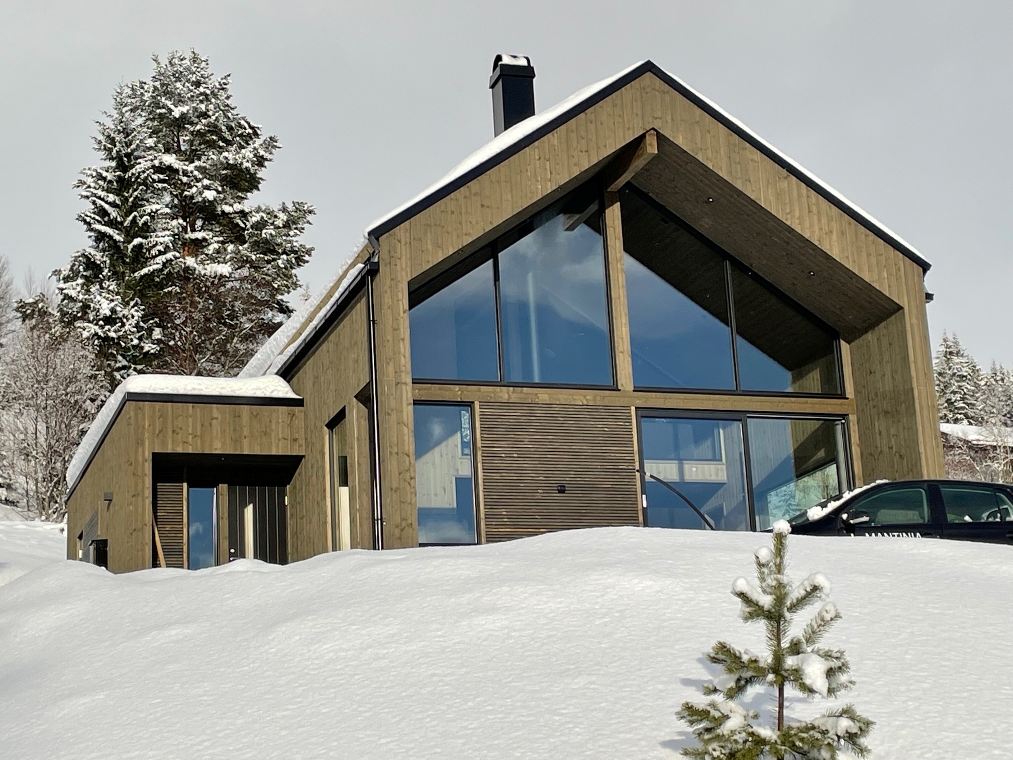 Modell B1 hytte vinterstid — snødekt landskap med hytte i mørkt trepanel og store vinduer